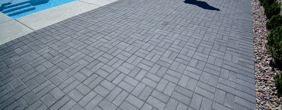 Paved patio at a home with grey stones, next to a pool and bushes.