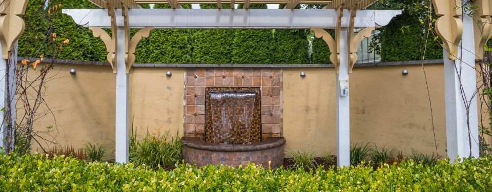 Stone water fountain underneath a pergolas in a lush backyard.